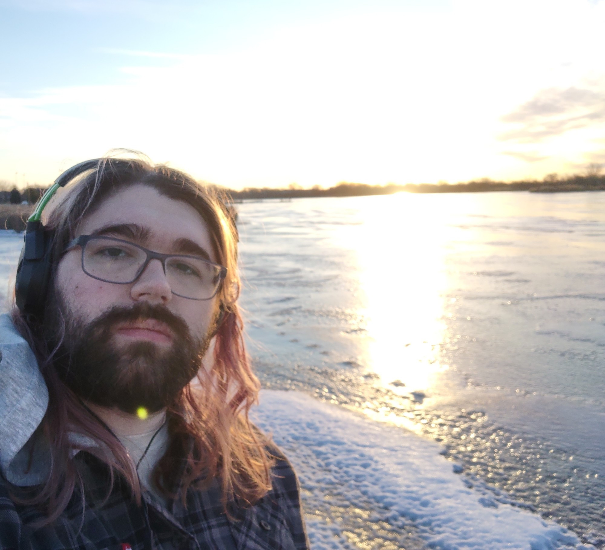 a selfie of a person standing next to an iced over lake, the sun making the sky a bright white, reflecting off of the ice. there are tracks across the ice as well.