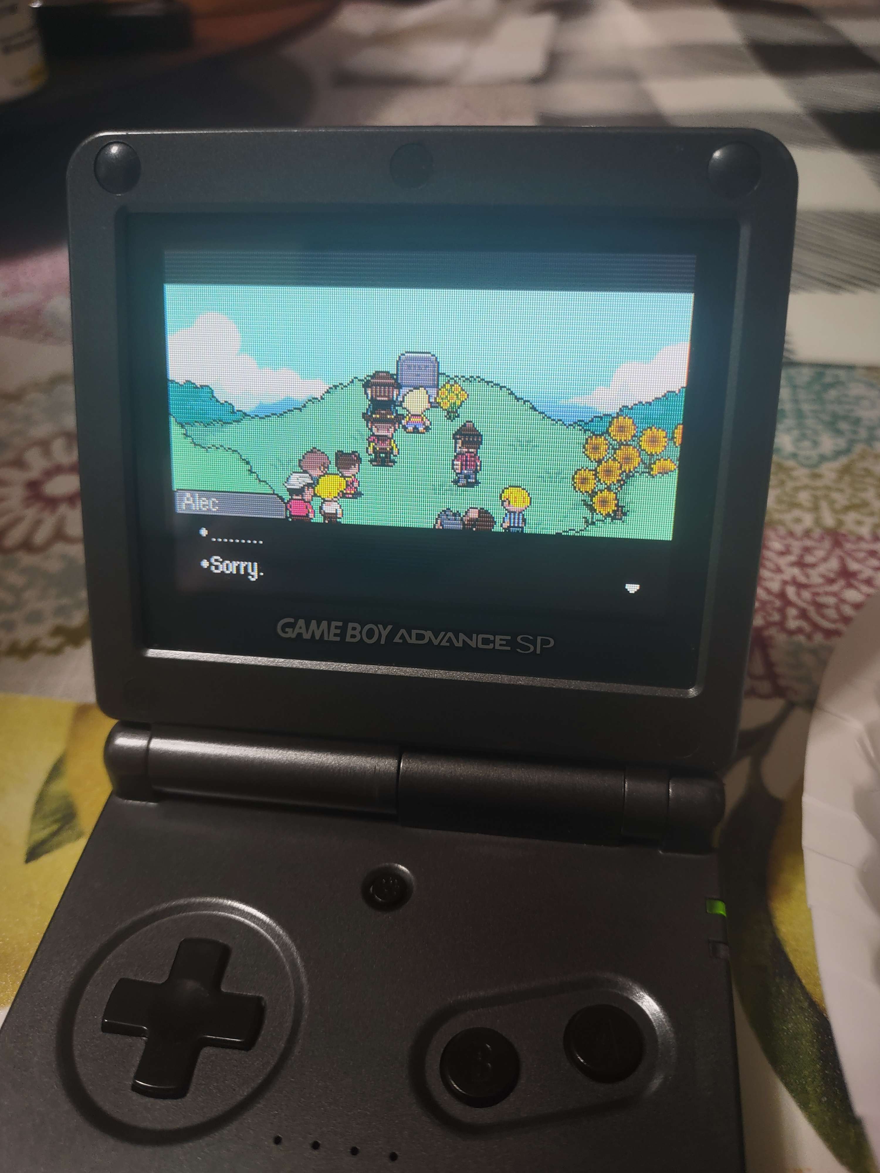 A photo taken of a Gameboy Advance SP playing the game Mother 3. The screen displays a group of characters around a gravestone, a textbox by a character named Alec saying sorry.