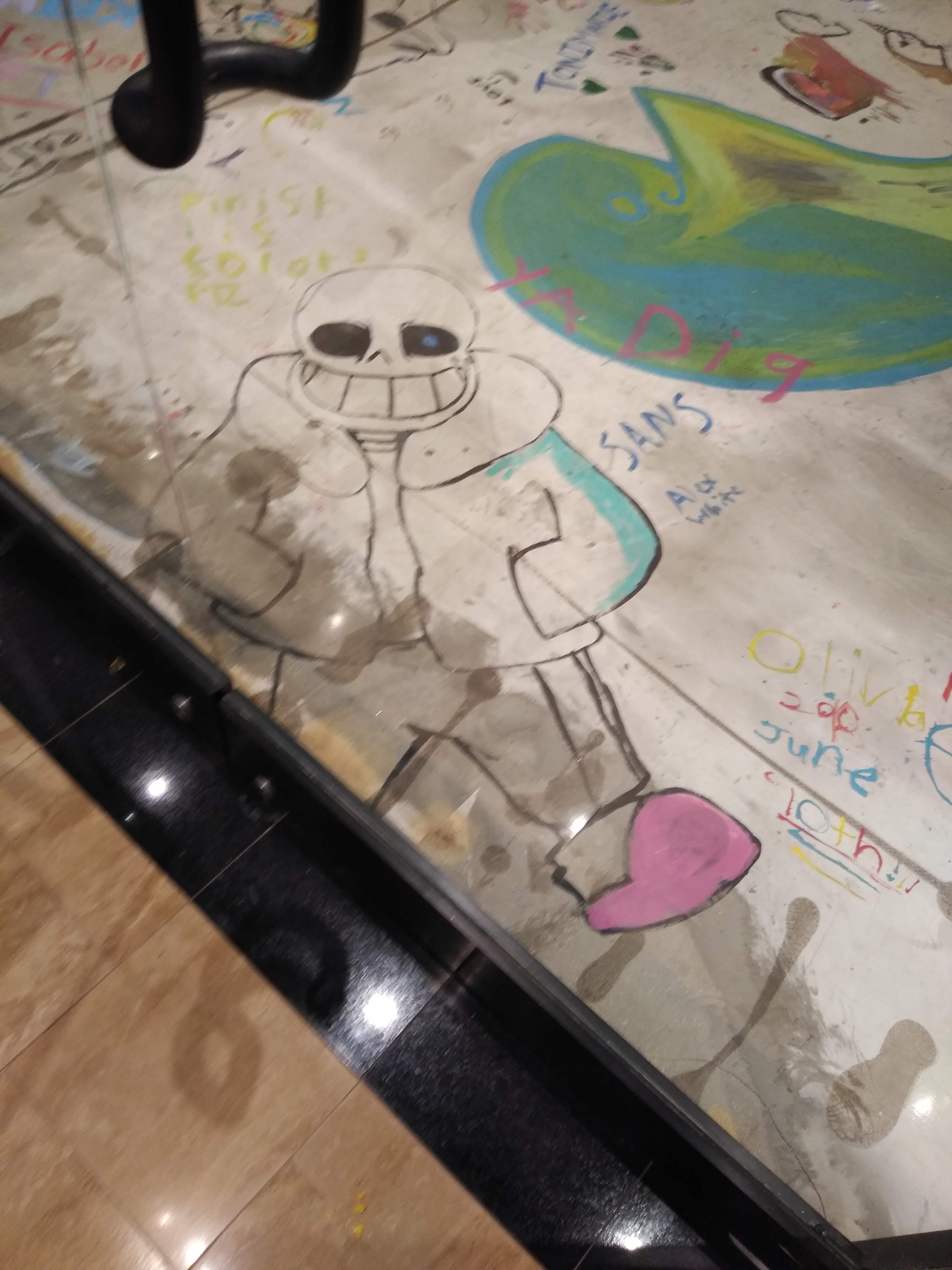 a photo taken behind a glass door, the ground being drawn on by many people, but the focus being on a large drawing of sans from the video game undertale.